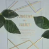 A clear, rectangular acrylic wedding invitation is displayed on a light gray surface, flanked by two artificial green leaves. The invitation features elegant gold text and a minimalist gold geometric design consisting of intersecting lines in the top right and bottom left corners.