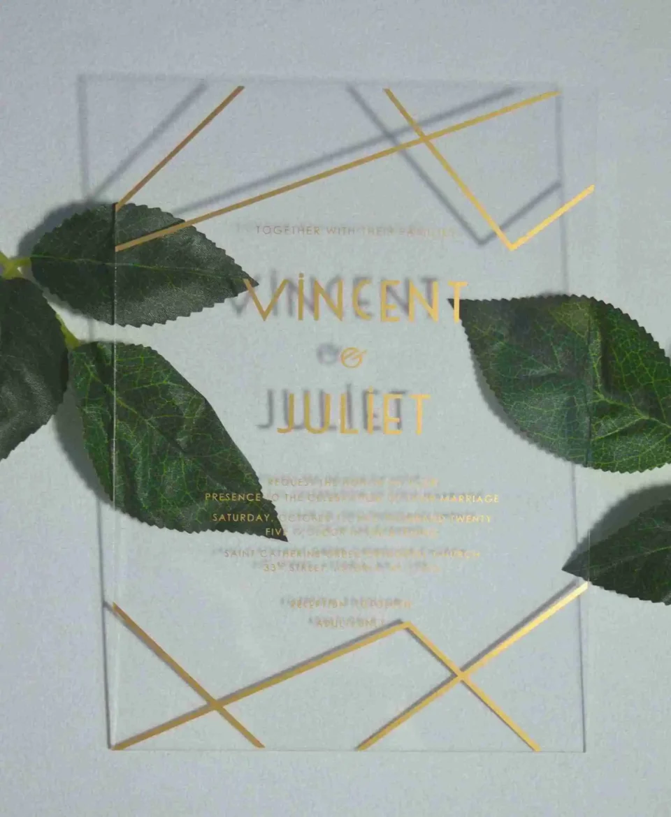 A clear, rectangular acrylic wedding invitation is displayed on a light gray surface, flanked by two artificial green leaves. The invitation features elegant gold text and a minimalist gold geometric design consisting of intersecting lines in the top right and bottom left corners.