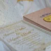A high-angle shot featuring an open peach-colored wedding invitation box set on a white surface with draped white fabric and a gold lace cuff. On the left, the closed lid of the box displays a central gold plaque with the monogram "R & P," framed by purple scrollwork accented with pearls. On the right, the open box base reveals a formal acrylic invitation for "James Parker & Karen Brett" printed with gold text. A gold RSVP card with matching scrollwork is partially tucked into a pocket in the open lid.