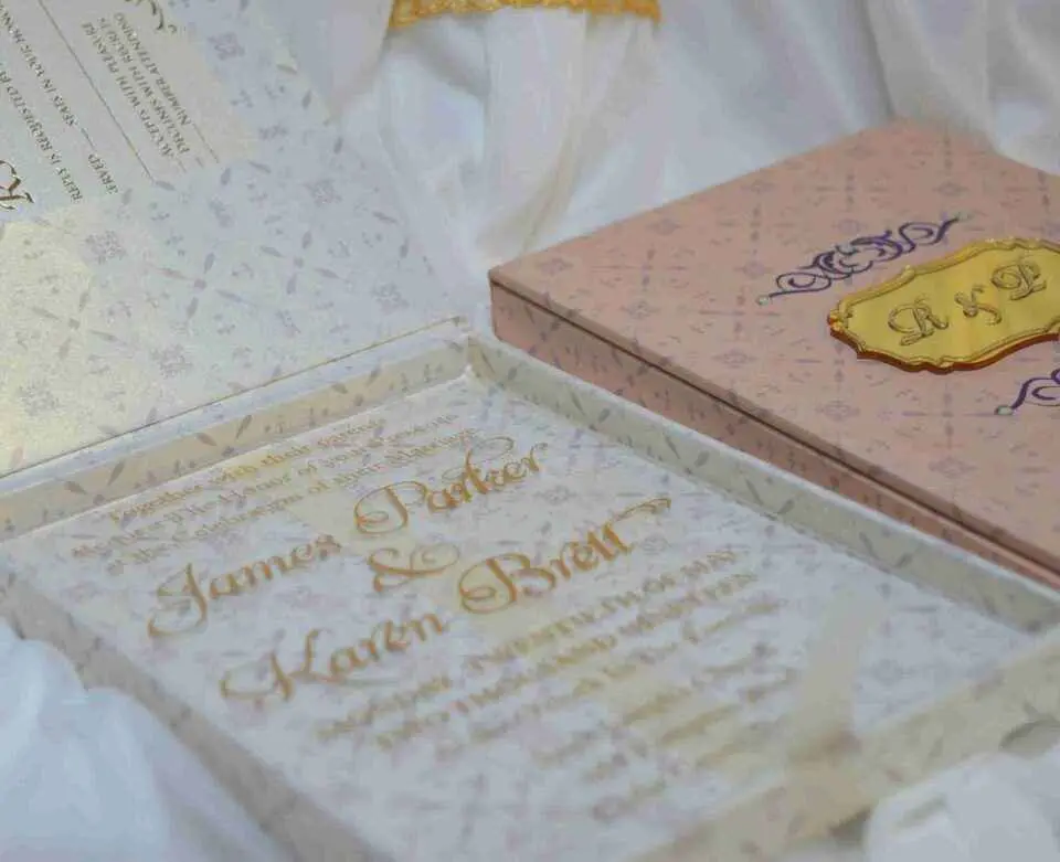 A high-angle shot featuring an open peach-colored wedding invitation box set on a white surface with draped white fabric and a gold lace cuff. On the left, the closed lid of the box displays a central gold plaque with the monogram "R & P," framed by purple scrollwork accented with pearls. On the right, the open box base reveals a formal acrylic invitation for "James Parker & Karen Brett" printed with gold text. A gold RSVP card with matching scrollwork is partially tucked into a pocket in the open lid.