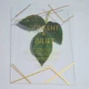A clear, rectangular acrylic wedding invitation is displayed on a light gray surface, flanked by artificial green leaves. The invitation features elegant gold text and a minimalist gold geometric design consisting of intersecting lines in the top right and bottom left corners.