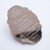 A silver mirror acrylic wedding invitation with a classic, ornate scalloped silhouette is shown at an angle against a white background. The highly reflective surface creates a liquid-like sheen, making the white printed text pop.