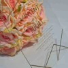 A transparent acrylic wedding invitation stands upright against a bouquet of peach-colored roses on a white surface. The invitation features elegant gold lettering and minimalist geometric gold lines in the corners.