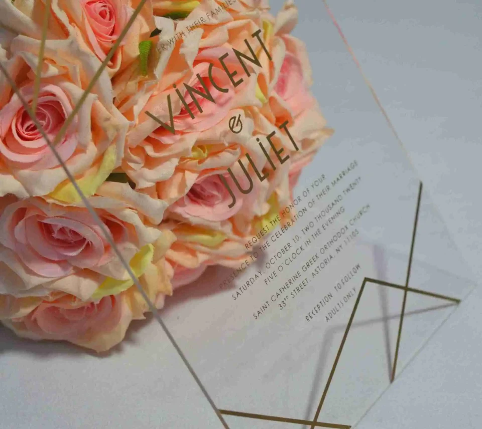 A transparent acrylic wedding invitation stands upright against a bouquet of peach-colored roses on a white surface. The invitation features elegant gold lettering and minimalist geometric gold lines in the corners.