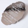 A silver mirror acrylic wedding invitation with a classic, ornate scalloped silhouette is shown at an angle against a white background. The highly reflective surface creates a liquid-like sheen, making the white printed text pop.
