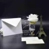 A sophisticated wedding invitation suite staged against a black background. The center of the display features a clear acrylic invitation card with white engraved text. The names are written in a large, elegant script. The event details below specify the date. To the left of the acrylic card is a white pointed-flap envelope. To the right is a small glass pedestal vase with a gold rim holding a large white rose, and a bronze-colored Eiffel Tower figurine. In the foreground, a cream-colored RSVP card and a small addressed envelope for are laid flat on the surface.