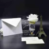 A sophisticated wedding invitation suite staged against a black background. The center of the display features a clear acrylic invitation card with white engraved text. The names are written in a large, elegant script. The event details below specify the date. To the left of the acrylic card is a white pointed-flap envelope. To the right is a small glass pedestal vase with a gold rim holding a large white rose, and a bronze-colored Eiffel Tower figurine. In the foreground, a cream-colored RSVP card and a small addressed envelope for are laid flat on the surface.