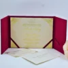 An open **magenta tri-fold wedding invitation folio** displayed against a white background. The central panel holds a cream-colored invitation card featuring: * **Gold foil script** for the names and the monogram at the top. * Ornate **gold foil filigree patterns** in each of the four corners. * Magenta silk ribbons securing the bottom corners of the card. The side panels feature magenta pockets. The left pocket contains a **cream RSVP card** with black decorative scrollwork, while the right pocket holds a coordinating card with printed text. A soft peach rose is partially visible in the lower right corner.