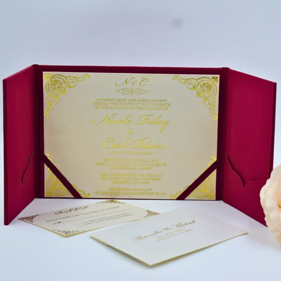An open **magenta tri-fold wedding invitation folio** displayed against a white background. The central panel holds a cream-colored invitation card featuring: * **Gold foil script** for the names and the monogram at the top. * Ornate **gold foil filigree patterns** in each of the four corners. * Magenta silk ribbons securing the bottom corners of the card. The side panels feature magenta pockets. The left pocket contains a **cream RSVP card** with black decorative scrollwork, while the right pocket holds a coordinating card with printed text. A soft peach rose is partially visible in the lower right corner.