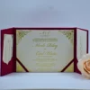 An open **magenta tri-fold wedding invitation folio** displayed against a white background. The central panel holds a cream-colored invitation card featuring: * **Gold foil script** for the names and the monogram at the top. * Ornate **gold foil filigree patterns** in each of the four corners. * Magenta silk ribbons securing the bottom corners of the card. The side panels feature magenta pockets. The left pocket contains a **cream RSVP card** with black decorative scrollwork, while the right pocket holds a coordinating card with printed text. A soft peach rose is partially visible in the lower right corner.