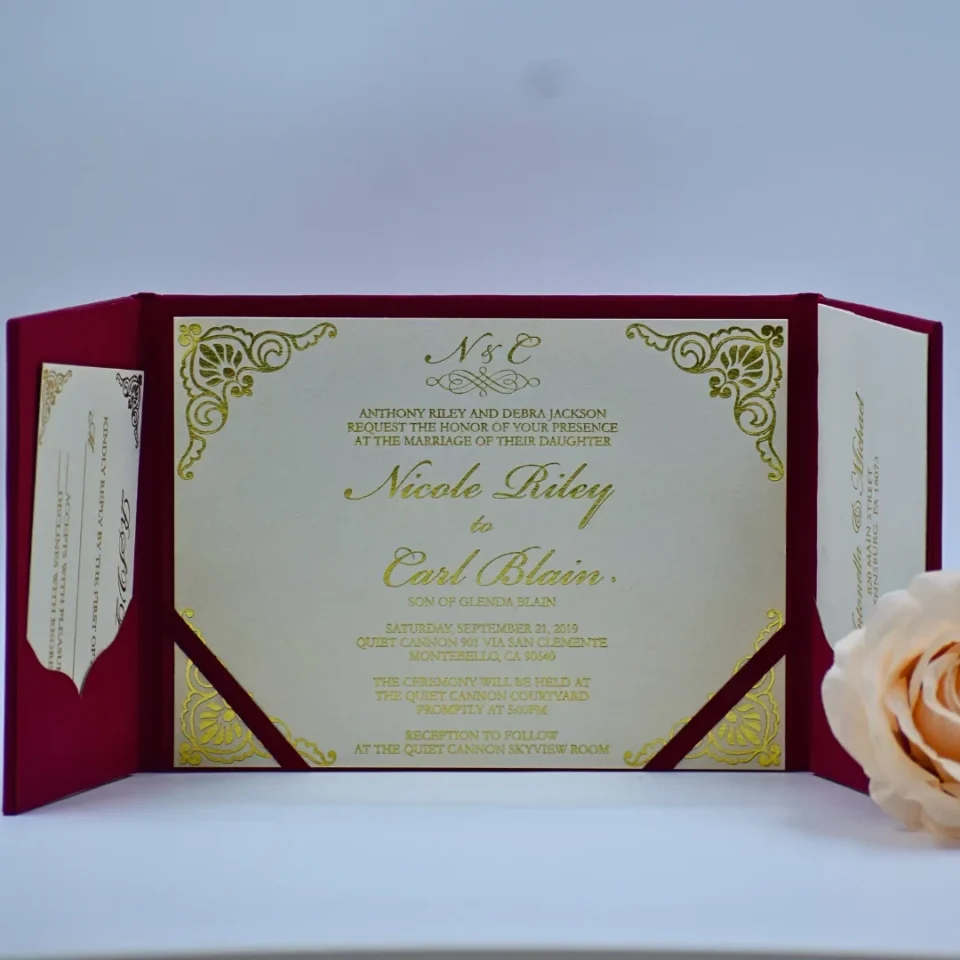 An open **magenta tri-fold wedding invitation folio** displayed against a white background. The central panel holds a cream-colored invitation card featuring: * **Gold foil script** for the names and the monogram at the top. * Ornate **gold foil filigree patterns** in each of the four corners. * Magenta silk ribbons securing the bottom corners of the card. The side panels feature magenta pockets. The left pocket contains a **cream RSVP card** with black decorative scrollwork, while the right pocket holds a coordinating card with printed text. A soft peach rose is partially visible in the lower right corner.