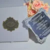 An open, white silk invitation box featuring a large, ornate gold monogram plaque on the left panel. Leaning against the right side is a clear acrylic wedding invitation with intricate silver-etched borders and white script lettering for the name. The invitation details a ceremony at the Hard Rock Hotel and Casino. In the background, soft peach-colored roses add a delicate floral accent against a neutral grey surface.
