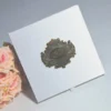 A top-down, slightly angled view of a square, white shimmering silk invitation box. In the center of the lid is a large, ornate gold metal plaque with an embossed oval crest containing stylized initials. A bouquet of soft peach and pink roses is visible in the lower-left corner, and the box sits on a smooth, light grey surface.