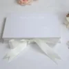 A top-down view of a rectangular, white silk wedding invitation box resting on a white fabric surface. The lid features a subtle, blind-embossed royal cres A wide, ivory satin ribbon is tied in a large bow at the bottom of the box. The scene is decorated with soft white and peach flowers and a gold Moroccan-style lantern in the background.