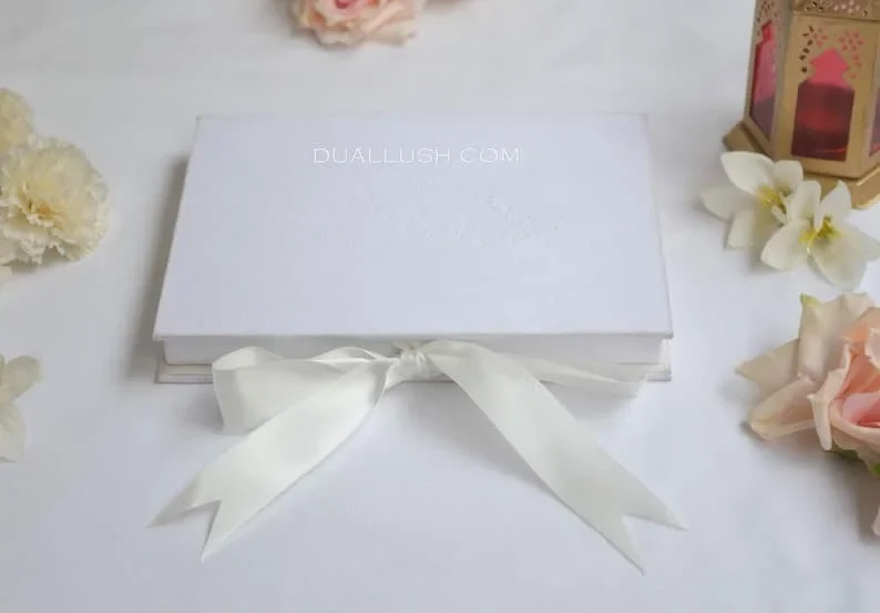 A top-down view of a rectangular, white silk wedding invitation box resting on a white fabric surface. The lid features a subtle, blind-embossed royal cres A wide, ivory satin ribbon is tied in a large bow at the bottom of the box. The scene is decorated with soft white and peach flowers and a gold Moroccan-style lantern in the background.