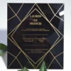 A square, elegant wedding invitation featuring a black acrylic or high-gloss finish with gold geometric line patterns and text. The invitation is held by a hand in the bottom-left corner against a plain white background, with green leaves resting over the .The visible in the soft-focus background on the left.