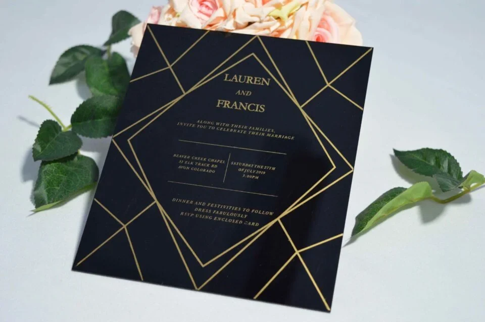 A square, elegant wedding invitation featuring a black acrylic or high-gloss finish with gold geometric line patterns and text. The invitation is held by a hand in the bottom-left corner against a plain white background, with green leaves resting over the .The visible in the soft-focus background on the left.A square, elegant wedding invitation featuring a black acrylic or high-gloss finish with gold geometric line patterns and text. The invitation is held by a hand in the bottom-left corner against a plain white background, with green leaves resting over the .The visible in the soft-focus background on the left.