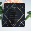 A square, elegant wedding invitation featuring a black acrylic or high-gloss finish with gold geometric line patterns and text. The invitation is held by a hand in the bottom-left corner against a plain white background, with green leaves resting over the .The visible in the soft-focus background on the left.