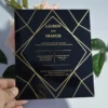 A square, elegant wedding invitation featuring a black acrylic or high-gloss finish with gold geometric line patterns and text. The invitation is held by a hand in the bottom-left corner against a plain white background, with green leaves resting over the .The visible in the soft-focus background on the left.