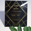 A square, elegant wedding invitation featuring a black acrylic or high-gloss finish with gold geometric line patterns and text. The invitation is held by a hand in the bottom-left corner against a plain white background, with green leaves resting over the .The visible in the soft-focus background on the left.