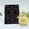 A modern wedding invitation made of glossy black cardstock or acrylic, featuring a striking geometric gold line pattern that creates a faceted, diamond-like frame around the central text. The invitation is staged on a white surface, flanked by soft white and pale yellow flowers. To the far left, a small portion of a bronze-colored Eiffel Tower figurine is visible.