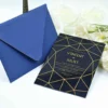 A wedding invitation suite displayed on a white surface, featuring a glossy black invitation card and a matching royal blue pointed-flap envelope. The card features a gold geometric line pattern that frames the central text. The gold text on the invitation.