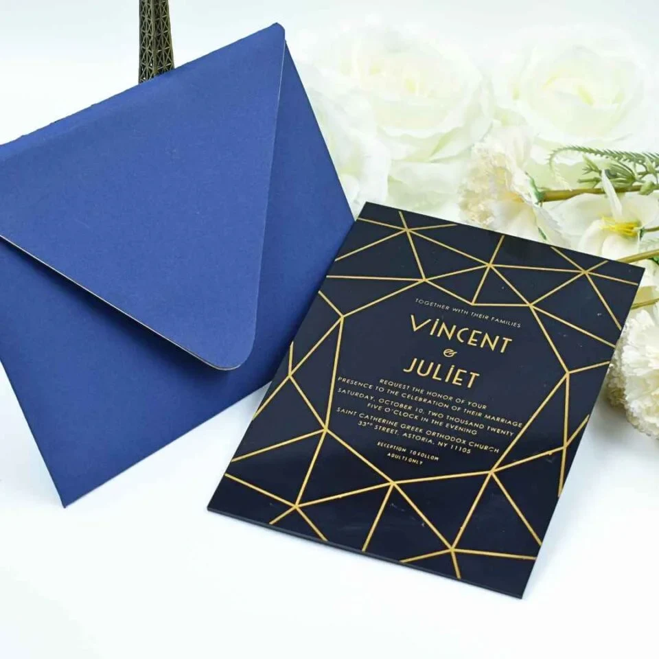 A wedding invitation suite displayed on a white surface, featuring a glossy black invitation card and a matching royal blue pointed-flap envelope. The card features a gold geometric line pattern that frames the central text. The gold text on the invitation.