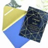 A wedding invitation suite displayed on a white surface, featuring a glossy black invitation card and a matching royal blue pointed-flap envelope. The card features a gold geometric line pattern that frames the central text. The gold text on the invitation.