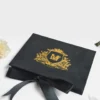 A high-angle view of an open **white luxury wedding invitation box** and its coordinating stationery. The inside lid features a gold-etched architectural illustration of a grand historic building. The suite includes: * A **clear acrylic invitation** with gold-foiled text and an intricate gold border, resting in a recessed compartment. * A **large ivory silk tassel** draped over the acrylic card. * An **RSVP card** with gold floral scrollwork in the corners. * An **Accommodations card** featuring elegant gold typography. * A white scalloped-edge envelope with a **gold foil monogram** on the flap. * Flowing **ivory satin ribbons** that weave through the arrangement on a clean white background.