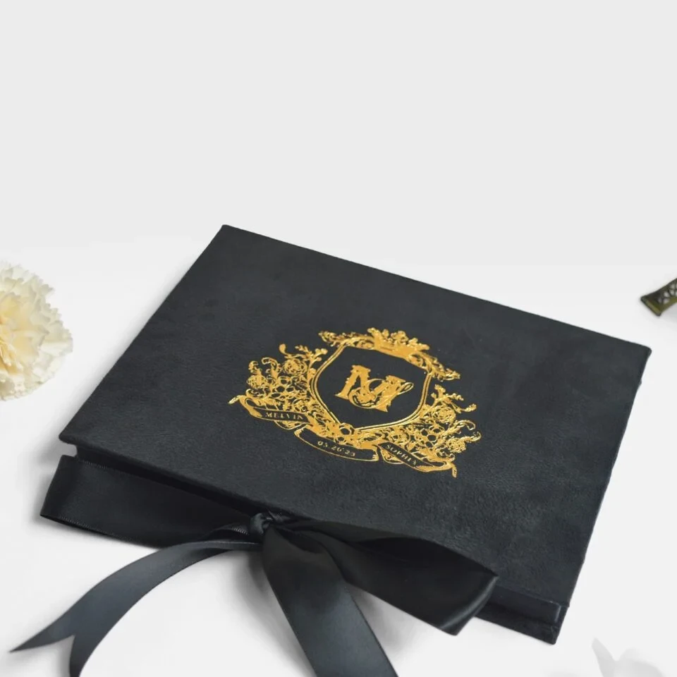 A high-angle view of an open **white luxury wedding invitation box** and its coordinating stationery. The inside lid features a gold-etched architectural illustration of a grand historic building. The suite includes: * A **clear acrylic invitation** with gold-foiled text and an intricate gold border, resting in a recessed compartment. * A **large ivory silk tassel** draped over the acrylic card. * An **RSVP card** with gold floral scrollwork in the corners. * An **Accommodations card** featuring elegant gold typography. * A white scalloped-edge envelope with a **gold foil monogram** on the flap. * Flowing **ivory satin ribbons** that weave through the arrangement on a clean white background.