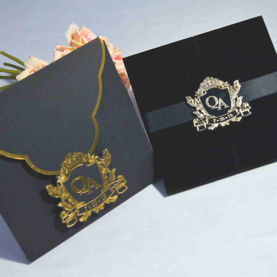 An overhead shot of a luxurious wedding invitation set. On the right is a **black velvet gatefold folio** held closed by a black satin ribbon and a polished **silver crest seal** featuring the monogram **"QA"** and the date **"7.20.19."** To the left is a matching **matte black envelope** with a gold-bordered curved flap, secured with an identical **gold crest seal**. A bouquet of soft peach roses is partially visible in the upper left corner against a plain white background.
