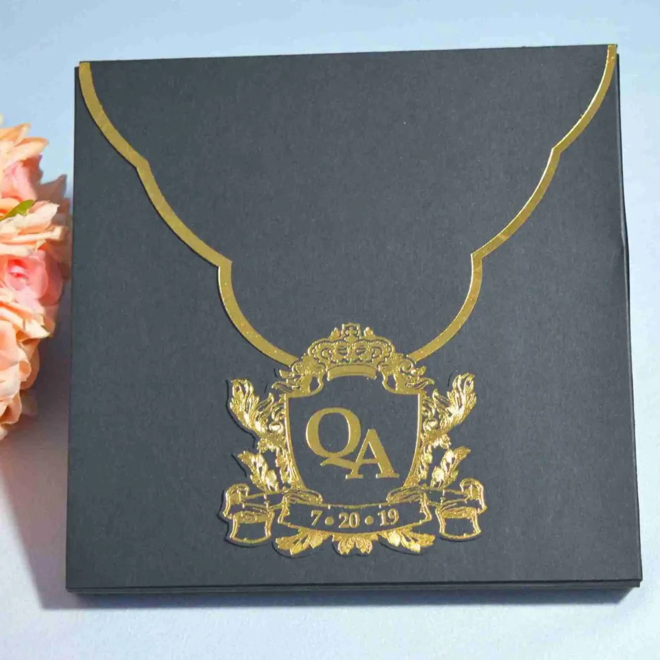 A top-down view of a **matte black square envelope** featuring a distinctively curved flap outlined with a thin **gold metallic border**. The envelope is secured by a large, ornate **gold crest seal** in the center, which showcases the monogram above the date in a matching metallic finish. To the left, a partial view of a **peach rose bouquet** provides a soft pop of color against the dark, formal stationery.