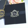 An overhead shot of a luxurious wedding invitation set. On the right is a **black velvet gatefold folio** held closed by a black satin ribbon and a polished **silver crest seal** featuring the monogram **"QA"** and the date **"7.20.19."** To the left is a matching **matte black envelope** with a gold-bordered curved flap, secured with an identical **gold crest seal**. A bouquet of soft peach roses is partially visible in the upper left corner against a plain white background.