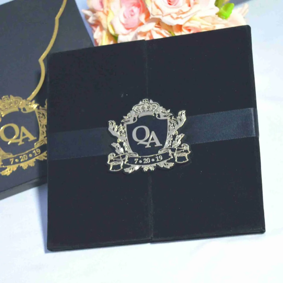 An overhead shot of a luxurious wedding invitation set. On the right is a **black velvet gatefold folio** held closed by a black satin ribbon and a polished **silver crest seal** featuring the monogram **"QA"** and the date **"7.20.19."** To the left is a matching **matte black envelope** with a gold-bordered curved flap, secured with an identical **gold crest seal**. A bouquet of soft peach roses is partially visible in the upper left corner against a plain white background.