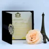 An open, luxurious **black velvet folio** showcasing a graduation invitation. The main invitation card is cream-colored with elegant **metallic gold** script and ornate yellow-gold floral details in the corners. The left side of the folio features a pocket containing smaller coordinated insert cards. In the foreground, a large **pale peach rose** and a miniature **bronze Eiffel Tower** figurine complete the sophisticated, Parisian-themed display.