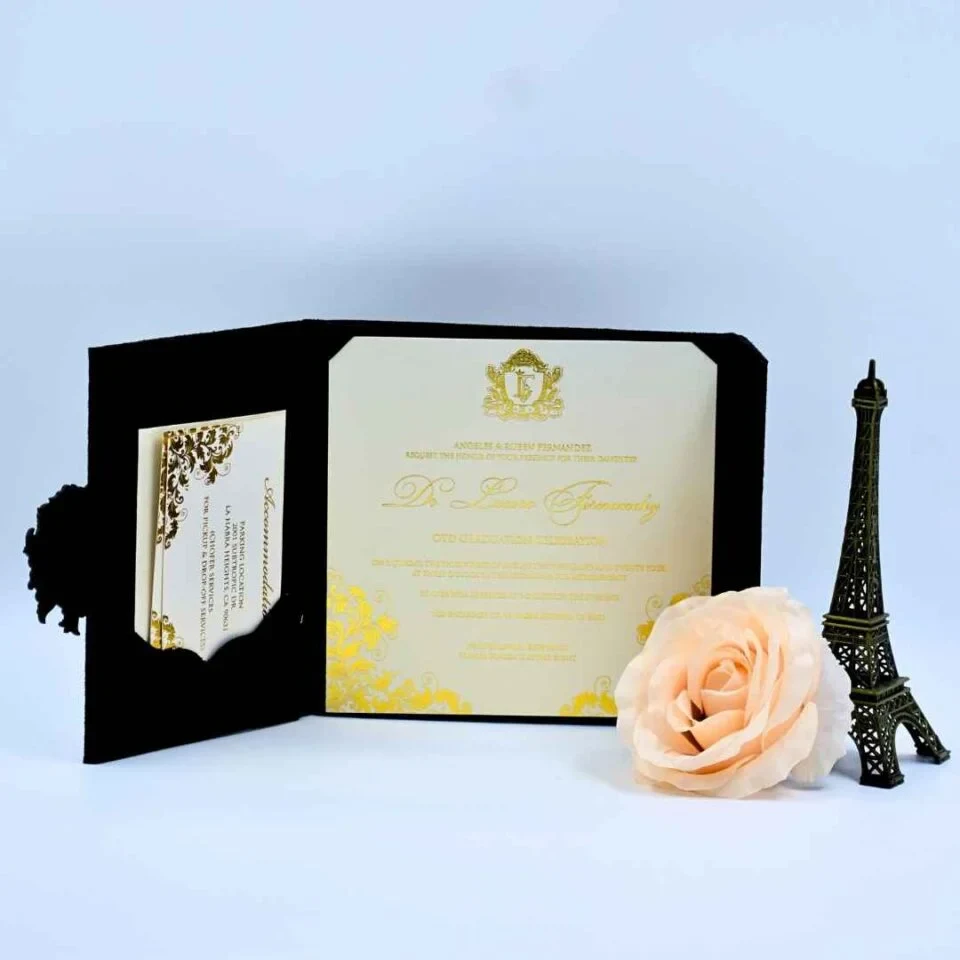 An open, luxurious **black velvet folio** showcasing a graduation invitation. The main invitation card is cream-colored with elegant **metallic gold** script and ornate yellow-gold floral details in the corners. The left side of the folio features a pocket containing smaller coordinated insert cards. In the foreground, a large **pale peach rose** and a miniature **bronze Eiffel Tower** figurine complete the sophisticated, Parisian-themed display.