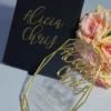 An elegant wedding invitation set featuring a black rectangular box and a geometric acrylic invitation. The black box on the left has written in gold cursive foil. To the right, a clear, geometric-shaped acrylic invitation rests against a bouquet of peach-colored roses. The acrylic piece also features the name in gold mirror acrylic script at the top, with the wedding details printed in smaller gold text below. The entire set is presented on a light blue surface.
