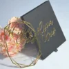 An elegant wedding invitation set featuring a black rectangular box and a geometric acrylic invitation. The black box on the left has written in gold cursive foil. To the right, a clear, geometric-shaped acrylic invitation rests against a bouquet of peach-colored roses. The acrylic piece also features the name in gold mirror acrylic script at the top, with the wedding details printed in smaller gold text below. The entire set is presented on a light blue surface.