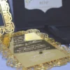 An open, black wedding invitation box showcased on a white fabric surface. The inner lid features a black pocket holding a cream-colored RSVP card decorated with gold floral scrollwork. The focal point is a stunning gold-mirrored acrylic invitation resting in the base of the box. The invitation is encased in an intricate, die-cut gold rose border that creates a frame around the elegant white-etched text. To the left, a portion of the box lid is visible, showing a matching gold-foiled rose pattern that coordinates with the invitation's ornate design.