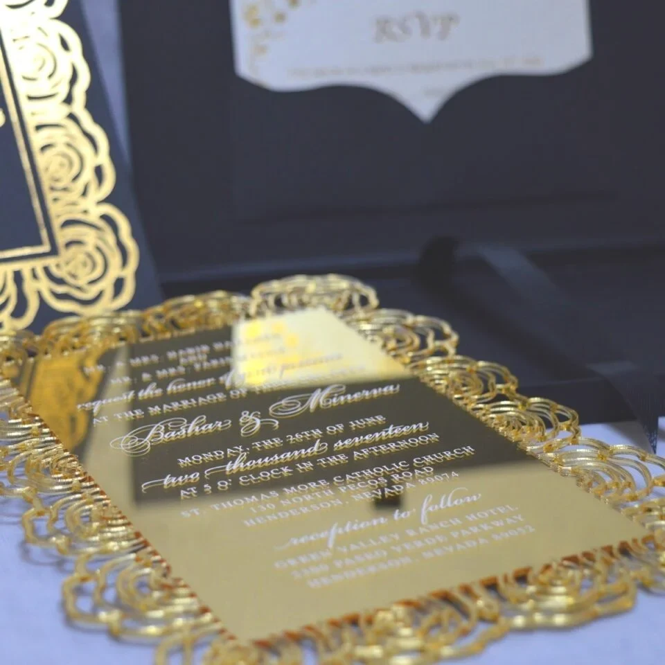 An open, black wedding invitation box showcased on a white fabric surface. The inner lid features a black pocket holding a cream-colored RSVP card decorated with gold floral scrollwork. The focal point is a stunning gold-mirrored acrylic invitation resting in the base of the box. The invitation is encased in an intricate, die-cut gold rose border that creates a frame around the elegant white-etched text. To the left, a portion of the box lid is visible, showing a matching gold-foiled rose pattern that coordinates with the invitation's ornate design.