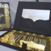 An open, black wedding invitation box showcased on a white fabric surface. The inner lid features a black pocket holding a cream-colored RSVP card decorated with gold floral scrollwork. The focal point is a stunning gold-mirrored acrylic invitation resting in the base of the box. The invitation is encased in an intricate, die-cut gold rose border that creates a frame around the elegant white-etched text. To the left, a portion of the box lid is visible, showing a matching gold-foiled rose pattern that coordinates with the invitation's ornate design.