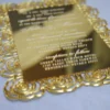 A close-up of a hand holding a rectangular gold-mirrored acrylic wedding invitation. The card is distinguished by a wide, die-cut border of intricate rose patterns that creates a decorative frame around the central text. The invitation features:Elegant white-etched typography announcing the marriage of Details for a ceremony date, location. A highly reflective surface that mirrors the person holding the camera, set against a blurred outdoor background.
