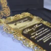An open, black wedding invitation box showcased on a white fabric surface. The inner lid features a black pocket holding a cream-colored RSVP card decorated with gold floral scrollwork. The focal point is a stunning gold-mirrored acrylic invitation resting in the base of the box. The invitation is encased in an intricate, die-cut gold rose border that creates a frame around the elegant white-etched text. To the left, a portion of the box lid is visible, showing a matching gold-foiled rose pattern that coordinates with the invitation's ornate design.