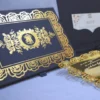 An open, black wedding invitation box showcased on a white fabric surface. The inner lid features a black pocket holding a cream-colored RSVP card decorated with gold floral scrollwork. The focal point is a stunning gold-mirrored acrylic invitation resting in the base of the box. The invitation is encased in an intricate, die-cut gold rose border that creates a frame around the elegant white-etched text. To the left, a portion of the box lid is visible, showing a matching gold-foiled rose pattern that coordinates with the invitation's ornate design.