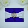 A luxurious royal blue silk gatefold invitation folder set against a clean white background with soft white roses. The folder features a high-shine, textured fabric and is secured by a wide white satin ribbon belly band. At the center of the ribbon is a large, ornate silver rhinestone brooch in a floral filigree design, adding a sparkling, regal touch. The folder is slightly ajar, revealing a hint of the printed invitation card inside.
