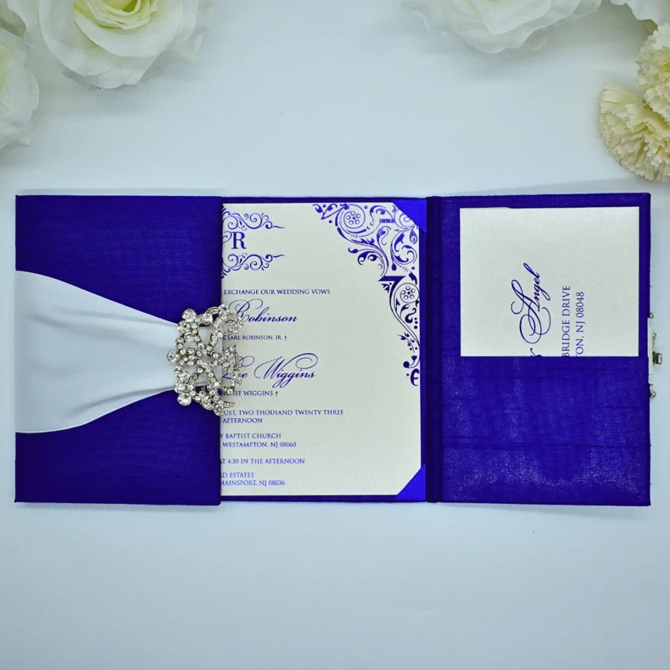 An open, three-panel **royal blue silk wedding invitation suite** displayed on a white surface with white floral accents. * **Left Panel:** Features a wide **white satin ribbon** gathered by a sparkling, ornate **silver rhinestone brooch**. * **Center Panel:** Holds the main invitation card, which is cream-colored with **royal blue script** and intricate blue scrollwork in the corners. The text mentions a wedding for **Robinson** and **Wiggins** in August 2023. * **Right Panel:** Includes a silk pocket containing a coordinating cream-colored response or detail card, also printed with blue ink. The overall design is vibrant and sophisticated, emphasizing a bold blue and crisp white color palette.