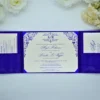 A fully opened **royal blue silk tri-fold invitation suite** laid flat on a white surface, framed by soft white and cream flowers at the top. * **Center Panel:** The main invitation card is cream-colored with **vibrant royal blue text** and ornate scrollwork in the corners. It announces the wedding of **Angel Robinson** and **Ronald Lee Wiggins** on Saturday, August 26, 2023. The card is held in place by blue silk corner ribbons. * **Left and Right Panels:** Each side features a blue silk pocket. The left pocket holds an **RSVP card**, and the right pocket holds an **additional details card**, both matching the cream and blue color scheme. The image provides a clear, top-down view of the entire wedding stationery set, highlighting the contrast between the deep blue silk and the elegant cream cardstock.