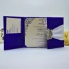 An open, three-panel **royal blue silk wedding invitation suite** displayed on a white surface with white floral accents. * **Left Panel:** Features a wide **white satin ribbon** gathered by a sparkling, ornate **silver rhinestone brooch**. * **Center Panel:** Holds the main invitation card, which is cream-colored with **royal blue script** and intricate blue scrollwork in the corners. The text mentions a wedding for **Robinson** and **Wiggins** in August 2023. * **Right Panel:** Includes a silk pocket containing a coordinating cream-colored response or detail card, also printed with blue ink. The overall design is vibrant and sophisticated, emphasizing a bold blue and crisp white color palette.