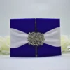 A luxurious royal blue silk gatefold invitation folder set against a clean white background with soft white roses. The folder features a high-shine, textured fabric and is secured by a wide white satin ribbon belly band. At the center of the ribbon is a large, ornate silver rhinestone brooch in a floral filigree design, adding a sparkling, regal touch. The folder is slightly ajar, revealing a hint of the printed invitation card inside.