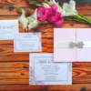 A complete blush pink and white wedding invitation suite arranged on a rustic wooden surface, accompanied by pink and white gladiolus flowers. The set includes: A closed gatefold folder: Made of pale pink silk or textured cardstock, secured with a white satin ribbon and a sparkling silver rhinestone brooch. Main invitation card: A white landscape card with pink floral corner illustrations and elegant script for the names. RSVP card: A matching smaller card with a response deadline meal choices. Return envelope: White with the couple's names and address printed in pink ink, featuring coordinating floral details.